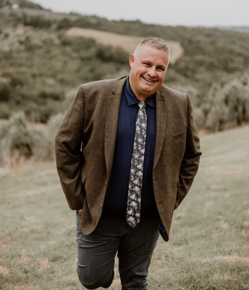 Josh Withers smiling outdoors in a brown jacket.